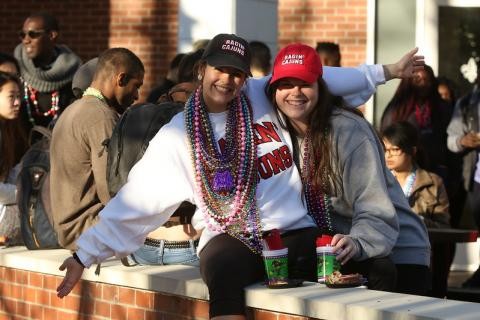 Two girls sitting on a brick wall on campus posing for the camera with mardi gras beads around their necks
