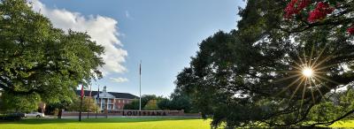 View through trees of UL Lafayette's Martin Hall, flags, and brick wall that says "University of Louisiana at Lafayette"