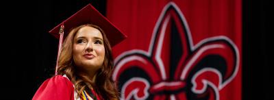 UL Lafayette honors student in full graduation attire standing in front of a fleur de lis.