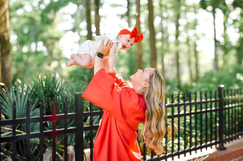 Online general studies grad holding her baby in the air near Cypress Lake. Grad wearing UL Lafayette cap and gown while baby wears a big red bow.