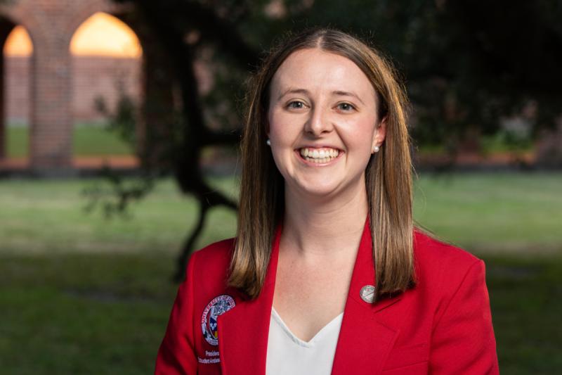 Portrait photograph of Mary McMahon smiling in front a campus building. She has on a red blazer with a University logo and a white blouse.