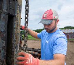 Online ms in engineering technology works on drilling equipment, wearing a blue shirt and red UL Lafayette cap.