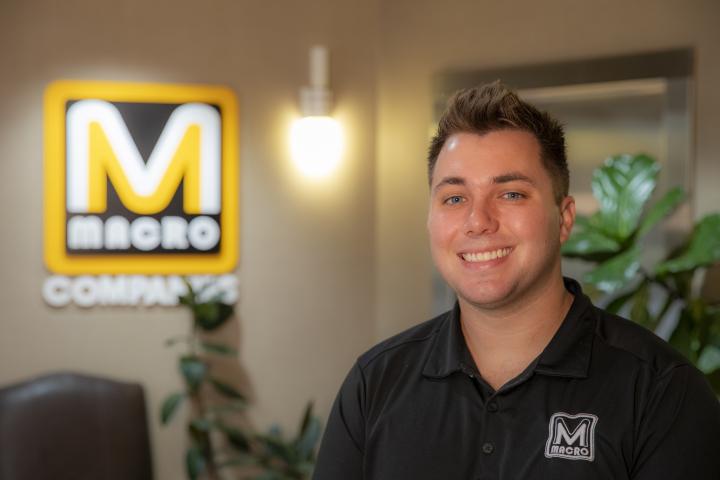 Caleb Robinson, BSBA in Management alum, is pictured indoors with a lit MACRO Companies sign behind him.