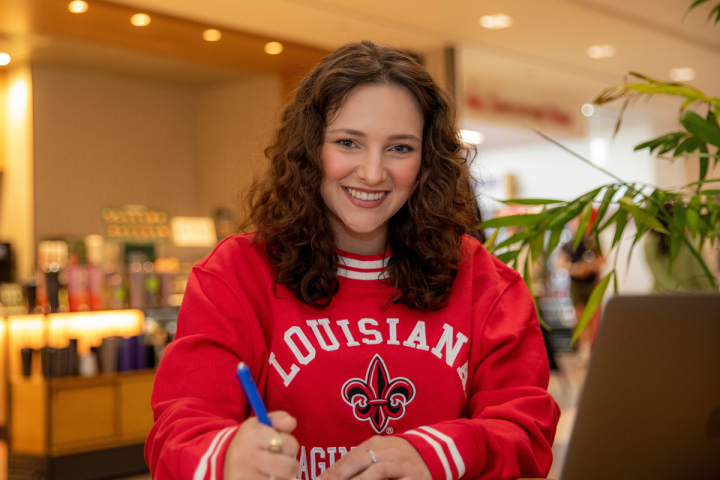 Brooke Perez, a student in the University of Louisiana at Lafayette's online criminal justice degree program, looks up at the camera and smiles as she works on her laptop in a coffee shop.