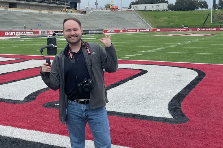 Daniel Richard, 2019 graduate of the University of Louisiana at Lafayette's marketing program, smiles for a picture on Cajun Field while capturing content with his phone.