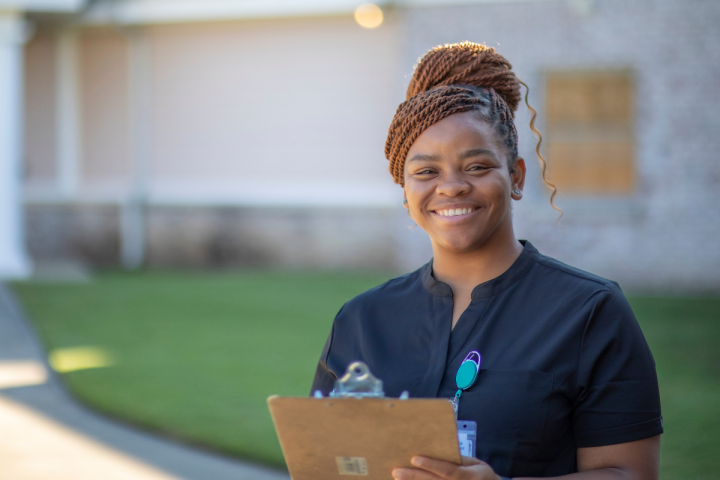 Destinee Francis, alumna of UL Lafayette's online bachelor's in health services administration program, smiles for a portrait in front of the facility she works in. She is wearing black scrubs and is holding a clipboard.