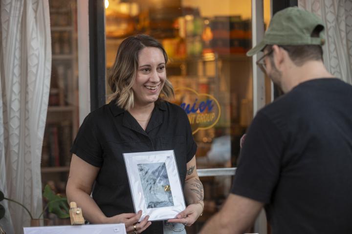 Lindsey Croall wears a black blouse and holds one of her watercolor illustrations while talking with a patron during an ArtWalk event.