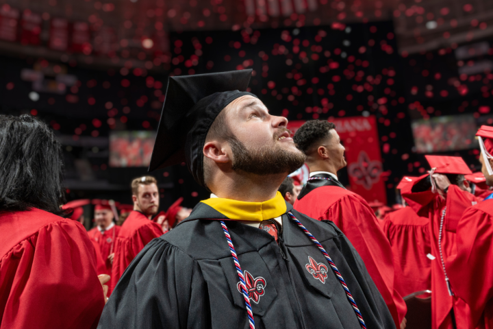 Online master's graduate wears a black cap and gown, gazing up as red confetti falls.