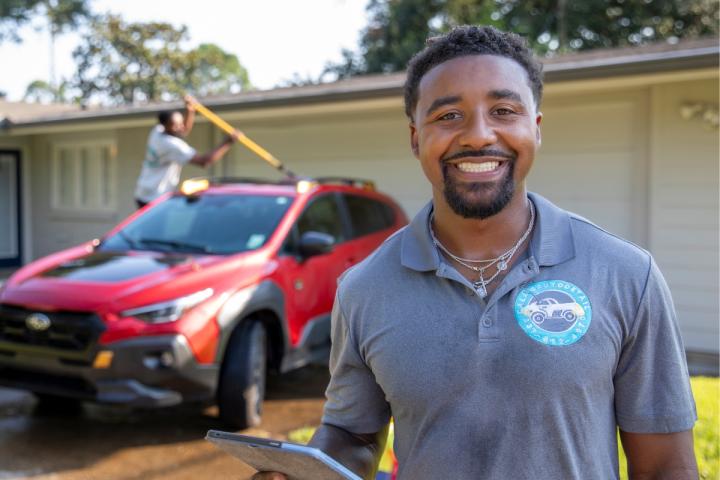 Entrepreneur Dillard Calais, graduate of UL Lafayette's online B.S.B.A. in Management program, smiles for a portrait while one of his employees details a client's car behind him.