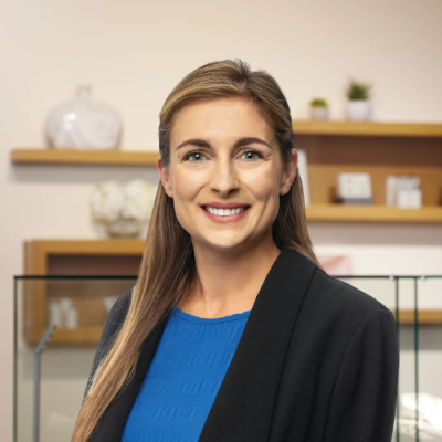 Jena Thompson smiles for headshot in an office setting, wearing a blue blouse and black blazer.