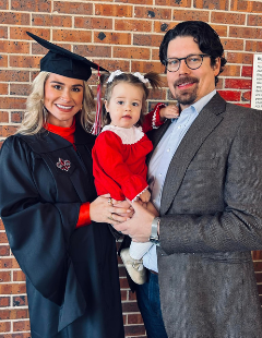 Online MBA grad Victoria Alleman smiles for photo with husband and daughter while wearing her master's regalia.
