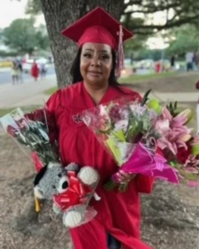 Chandra Cousin smiles wearing a red UL Lafayette cap and gown, holding gifts and bouquets after commencement ceremonies.