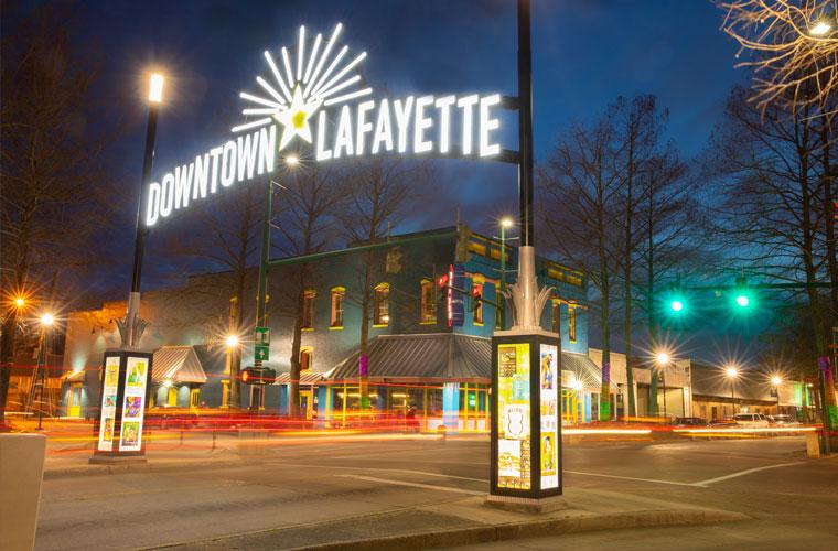 Nighttime shot of the neon Downtown Lafayette sign lit up while traffic passes underneath