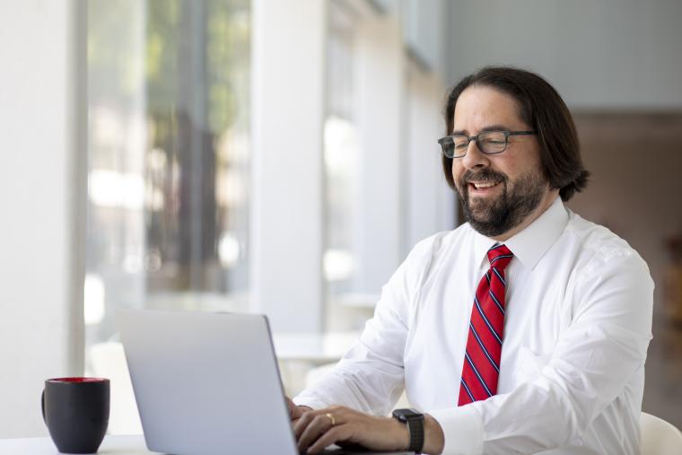 Josh Dufrene smiles while working on laptop, wearing a white collared shirt and a red tie.