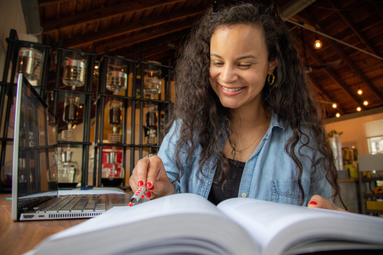 Nursing student sits in a coffee shop smiling over a textbook, wearing a black shirt and denim jacket.