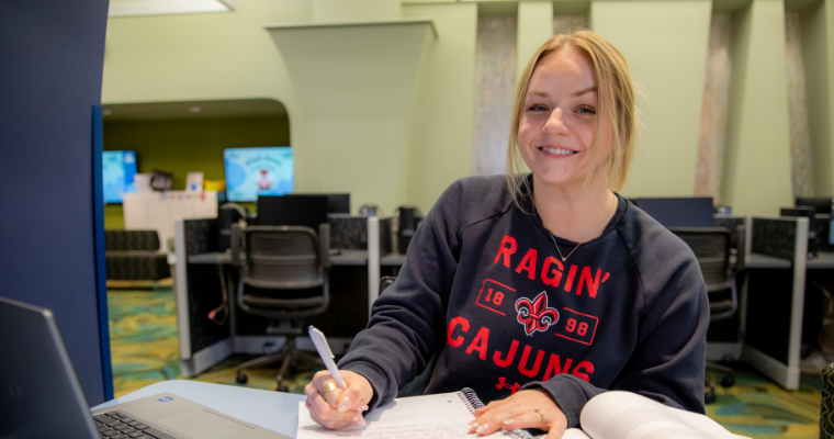 A young woman wearing a Ragin' Cajuns sweatshirt smiles as she works on her laptop and writes in her notebook at the public library.