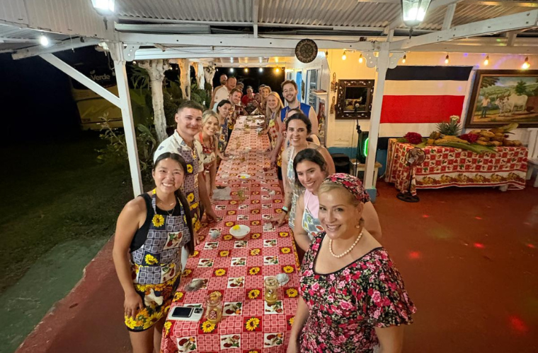MBA students studying abroad stand around a long table before eating their dinner together.