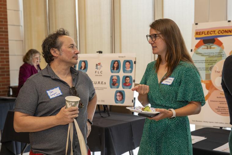 Two faculty members, a man and a woman, are pictured in discussion with research poster presentations in the background.