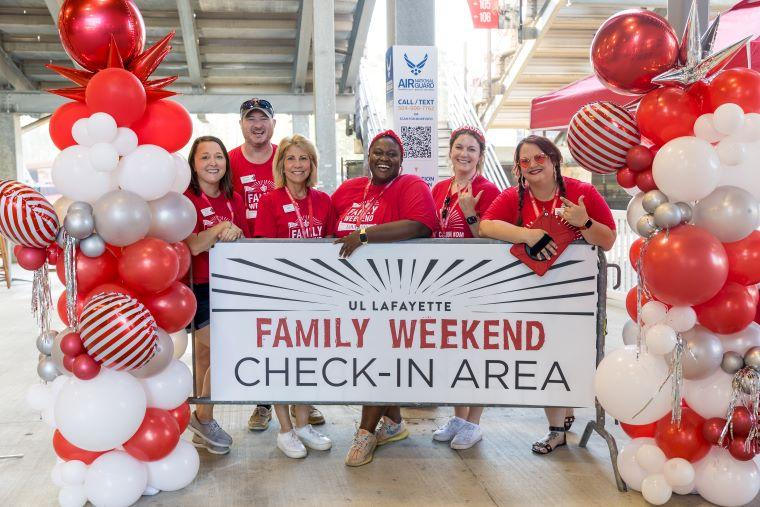 Family Weekend staff pose in front of a display that reads "UL Lafayette Family Weekend Check-In Area"
