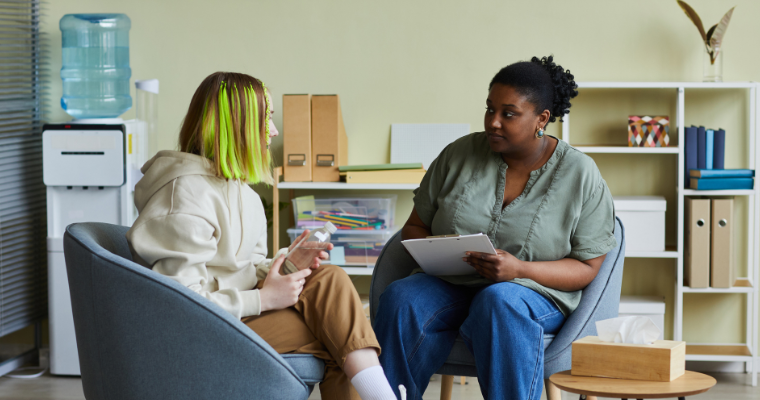 A social worker wearing jeans and a green blouse speaks with a young woman wearing a hoodie with green streaks in her hair.
