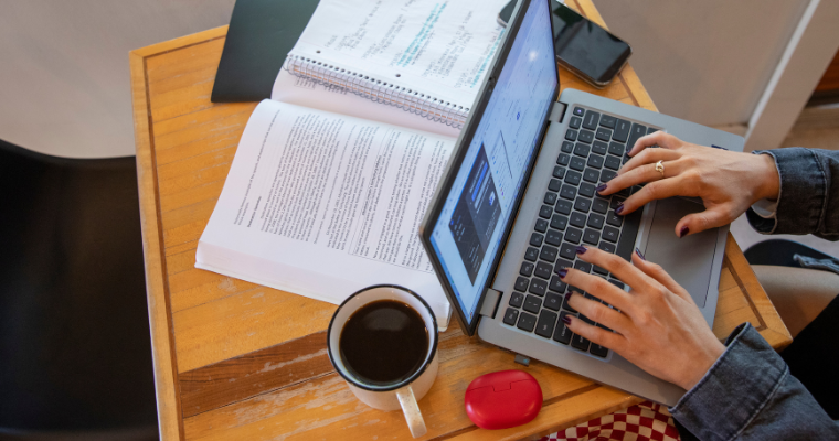A UL Lafayette online student types on laptop with a cup of black coffee and homework on the table.