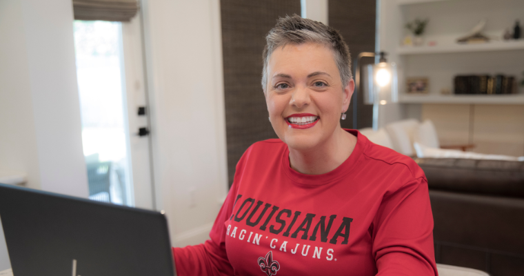 UL Lafayette alumni smiles in a red Ragin' Cajun t-shirt while working on a laptop, balancing online school and work.