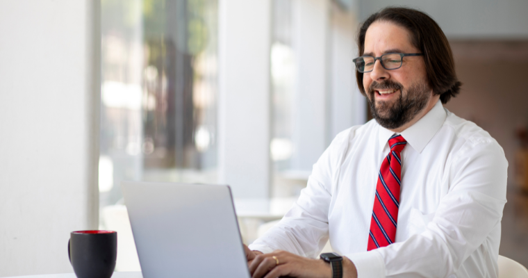 Professional wears a button down and tie, smiling while completing online MBA in HR homework on a laptop.