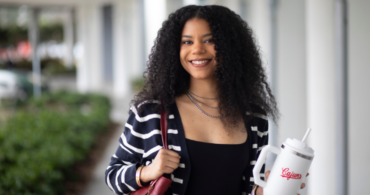 Online MBA in Sales Leadership student smiles while carrying a work tote bag and a UL Lafayette "Cajuns" cup.