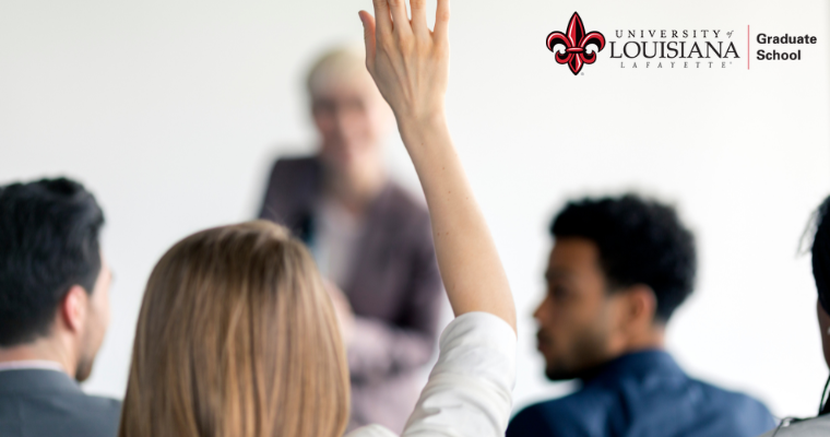 a view from behind a sitting adult student raising her hand to ask the teacher int eh front a question