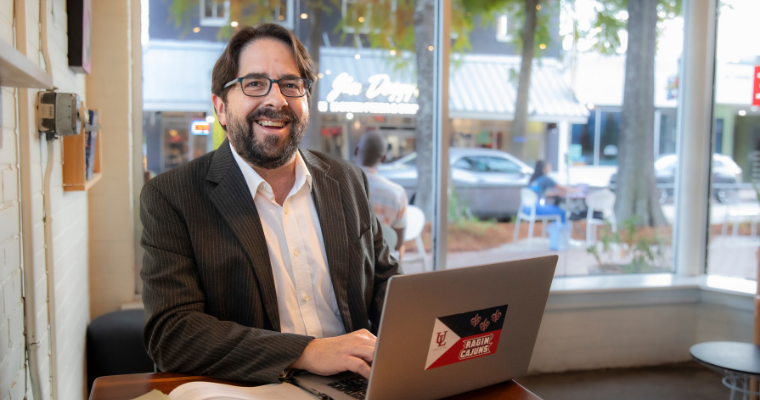 UL Lafayette online advisor smiles while working on laptop in a coffee shop.