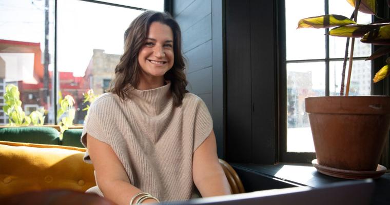 Online student smiles as she works on a laptop in a coffee shop. She is wearing a beige sleeveless sweater.