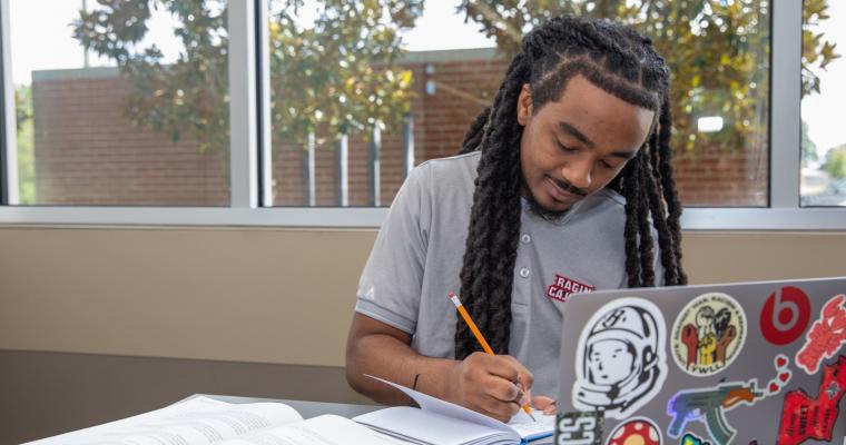 Student in UL Lafayette's online degree program studies at his laptop in a private library study room. He is wearing a gray University polo.