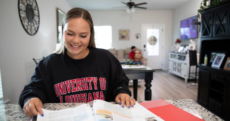Student in the online general studies degree program smiles while flipping through a textbook at home. She's wearing a black UL Lafayette sweatshirt.