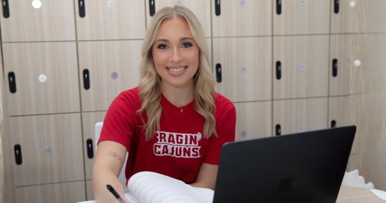 Online student studies at laptop in front of a wall of lockers. The student is smiling and wearing a red Ragin' Cajuns t-shirt.