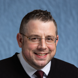 A man with rectangular glasses grins at the camera, he has vaguely curly hair and a dimple in his chin.
