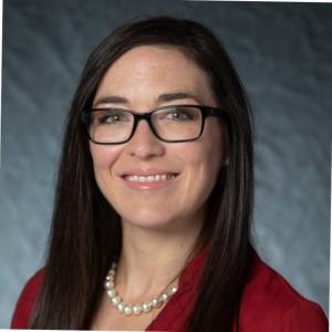 Portrait photograph of Camille Breaux smiling wearing pearl, glasses, and a red blazer.