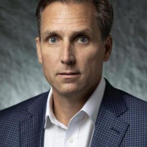 Portrait photo of Dr. Brian Bolton in a checkerboard navy blue suit and a white button down dress shirt underneath with a grayscale backdrop.
