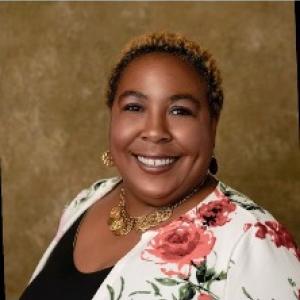 Portrait photograph of Dr. Dione Davis smiling she is wearing gold earrings, a gold necklace, a white shawl with roses designed on it, and a black top with a gold backdrop..