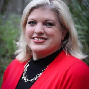 Portrait photo of Mary Baudoin smiling wearing a black turtleneck blouse and a red blazer.