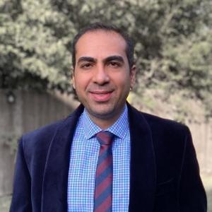 Dr. Farzad Ferdowsi faces the camera and smiles. He is wearing a suit and standing outdoors in front of a tree. 