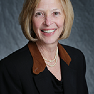 Portrait photograph of Lucy Henke smiling and wearing pearls, a black dress, and a black blazer with a brown collar.
