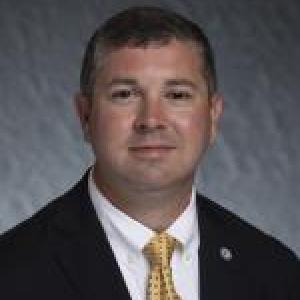 Portrait photograph of Jeremy T. Navarre smiling wearing a white button down shirt with a gold tie and a black blazer.