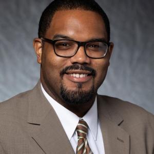 Portrait photograph of Kevin Guillory smiling and wearing glasses, a white button down shirt, a brown tie, and a brown blazer.