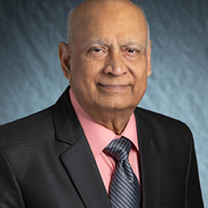 Portrait photo of Spuma Rao smiling and wearing a salmon button down shirt, spiral designed tie, and a black blazer.