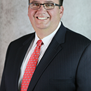 Portrait photograph of Geoff Steward smiling and wearing glasses, a white button down shirt, a red tie, and a black blazer.