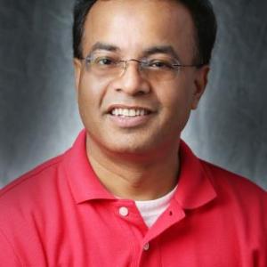 Portrait photograph of Ram Thakur smiling and wearing glasses and a red polo shirt.