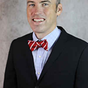 Portrait photograph of Linus Wilson smiling and wearing a blue button down shirt with a bowtie and a blue blazer.