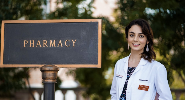 Bachelor of General Studies alumni Kailee Marikar stands in a white lab coat near a "pharmacy" sign shortly before earning her doctor of pharmacy degree.