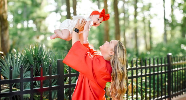 Online general studies grad holding her baby in the air near Cypress Lake. Grad wearing UL Lafayette cap and gown while baby wears a big red bow.
