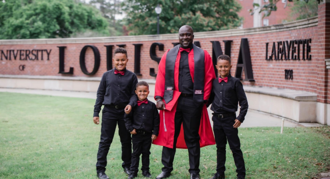 General Studies graduate and his 3 sons smile in front of the University of Louisiana at Lafayette sign in formal attire
